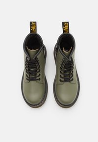 Botines de piel verde con cordones negros, puntera redonda, detalles de costura y una tira de tirón. Presentan una suela de goma marrón resistente.