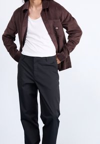 Chemise en velours côtelé brun, débardeur blanc et pantalon noir. La chemise présente un design à boutonnière et une poche avec un marquage.