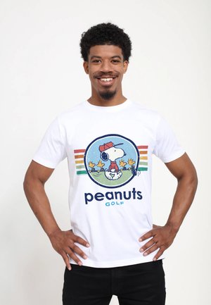 Uśmiechnięty mężczyzna z kręconymi włosami, ubrany w białą koszulkę z wizerunkami Snoopy'ego i Woodstocka grających w golfa, z napisem „peanuts golf” pod wzorem.