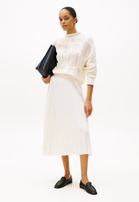 Hvit strikket genser med teksturerte mønstre, kombinert med en plissert hvit midiskjørt. Svart clutch-veske og svarte loafers fullfører antrekket.
