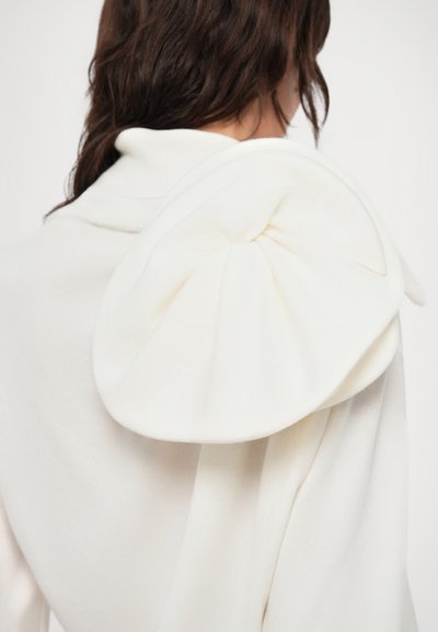 Balts, pildīts džemperis ar lielu, strukturētu loku detaļu uz pleca, izgatavots no mīkstas, teksturētas auduma.