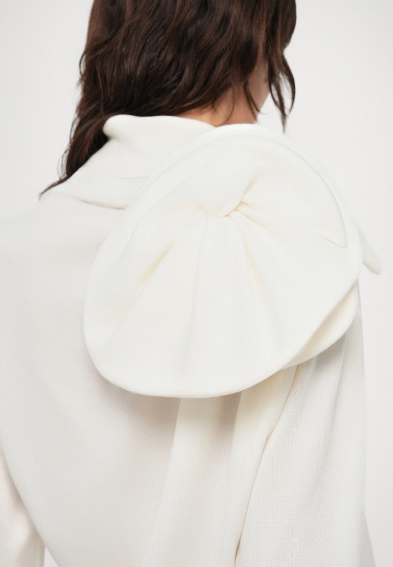 Maglione bianco imbottito caratterizzato da un grande dettaglio a fiocco strutturato sulla spalla, realizzato in un tessuto morbido e testurizzato.