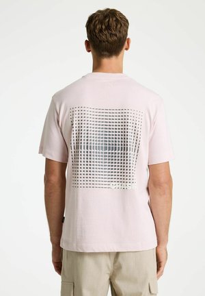 Man die lichtroze T-shirt met korte mouwen draagt met zwart- en grijsgeblokt patroon op de achterkant, beige cargobroek, staand met de rug naar de camera.