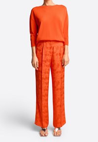 Oranje gebreide trui met driekwart mouwen, gecombineerd met oranje wijde broek met een textuur in bloempatroon en zilveren sandalen.