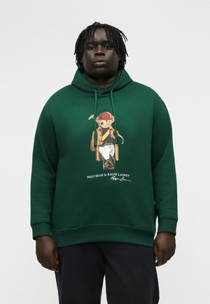 Άνδρας που φοράει σκούρο πράσινο φούτερ με κουκούλα, με γραφικό ενός αρκούδου ντυμένου με πόλο που κρατάει ένα σφυρί και το κείμενο "POLO BEAR by RALPH LAUREN".