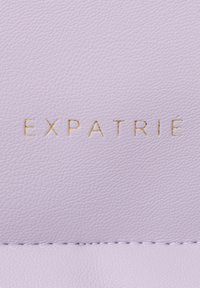 Cuoio di lavanda con una superficie texturizzata, caratterizzato da un testo dorato embosso "EXPATRIÉ" centrato sul materiale e una cucitura alla base.