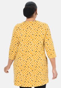 Mosterdgele tuniek met een stippenpatroon in het wit en zwart. Ontworpen met driekwartmouwen en zijsplitten voor meer bewegingsvrijheid.