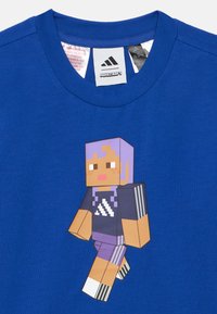 Kék póló, melynek elején egy Minecraft-stílusú karakter látható, aki sötét ruhát visel fehér csíkokkal és lila hajjal.