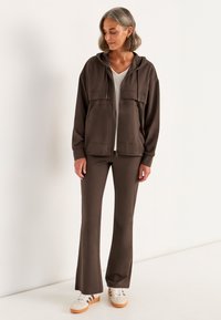 Brun zip-up hættejakke med to forlommer, parret med matchende brune flared bukser, båret med hvide sneakers med grå detaljer.
