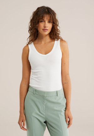 Frau mit lockigem braunem Haar, die ein weißes ärmelloses Oberteil und hellgrüne, maßgeschneiderte Hosen trägt und vor einem beigen Hintergrund steht.