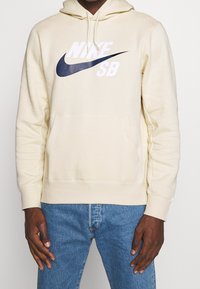 Man i beiga Nike SB-hoodie och blå jeans, stående med avslappnade händer vid sidorna mot en enfärgad bakgrund.