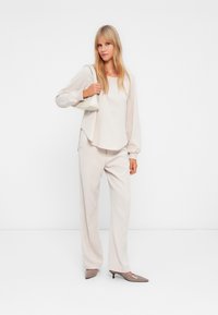 Μπεζ μπλούζα με μακριά μανίκια και χαλαρή εφαρμογή, συνδυασμένη με ασορτί παντελόνι σε φαρδιά γραμμή και ανοιχτό γκρι γόβες; κρατώντας μια μικρή λευκή τσάντα.