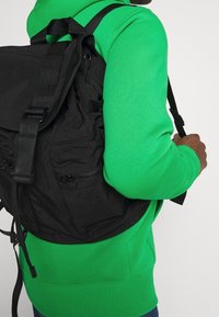Zaino nero in nylon con finitura testurizzata, spallacci regolabili e scomparti con zip, indossato sopra un maglione verde brillante.