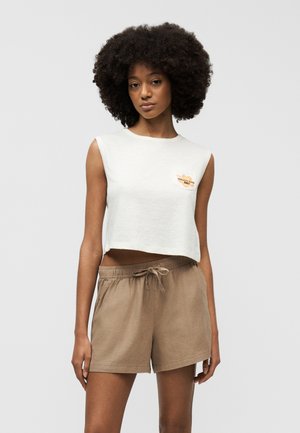 Mladá žena s prirodzenými kučeravými vlasmi, oblečená v bielom krátkom tielku bez rukávov s logom a béžových šortkách so sťahovacou šnúrkou, stojaca pred jednoduchým pozadím.