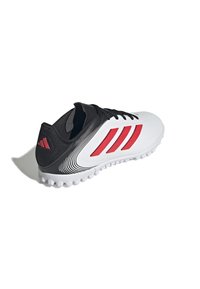 Zapatilla deportiva blanca con acentos negros, que cuenta con tres rayas rojas, una superficie texturizada y una suela de goma moldeada con clavos redondeados.