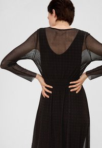Doorzichtig zwart jurkje met lange mouwen, V-hals aan de achterkant en een gerimpelde taille. Bevat een subtiele patroonstructuur door het geheel, voortvloedend naar een lange zoom.