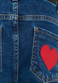 Jeans in denim di colore blu scuro, con una vistosa toppa a forma di cuore rosso sulla tasca posteriore e cuciture gialle a contrasto.