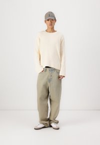 Kremfarget ribbestrikket genser, lysegrønne baggy jeans og grå caps. Modellen har på seg hvite sneakers med grå og brune detaljer. Enkelt design.