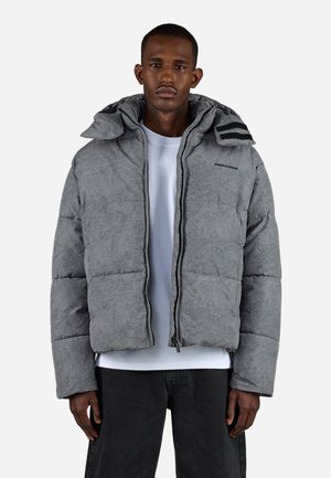 Man wearing a gray padded jacket with a hood over a white shirt and black pants, standing against a plain white background.