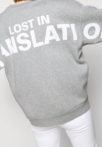Persona vistiendo una sudadera gris con texto blanco parcialmente visible "LOST IN ISLATION" y pantalones blancos, vista desde atrás.