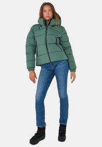 Zielona kurtka puchowa z kapturem, zapinana na zamek z przodu i dwoma bocznymi kieszeniami. Lekka, pikowana konstrukcja z elastycznymi mankietami. Połączona z niebieskimi dżinsami i czarnymi butami.
