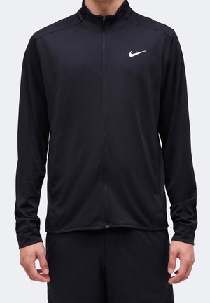 Homme portant une veste de sport Nike noire à fermeture éclair intégrale avec un logo Nike blanc sur la poitrine gauche, debout devant un fond uni.