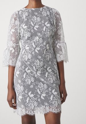 Robe en dentelle florale blanche avec manches cloche 3/4 et ourlet festonné portée par une personne sur fond uni.