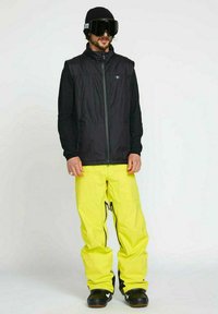 Schwarze isolierte Weste über einem schwarzen Langarmoberteil, kombiniert mit leuchtend gelben Schneehosen. Eine schwarze Mütze und große schwarze Skibrillen runden das Outfit ab.