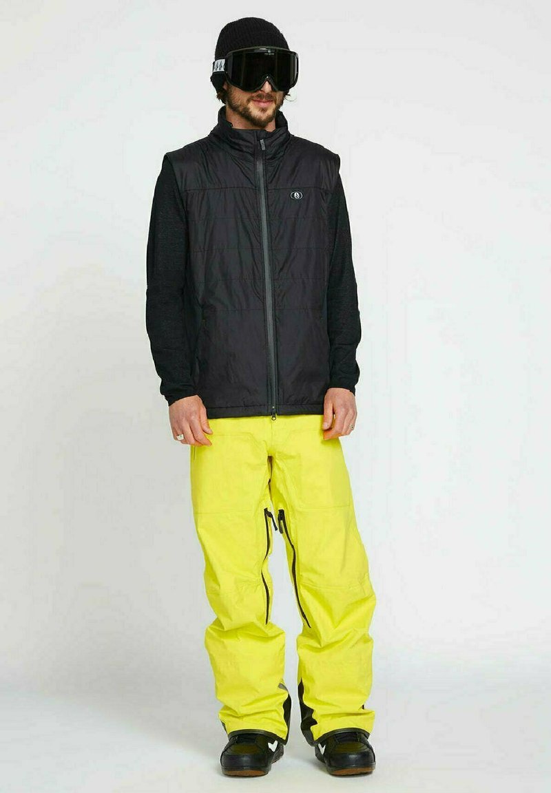 Schwarze isolierte Weste über einem schwarzen Langarmoberteil, kombiniert mit leuchtend gelben Schneehosen. Eine schwarze Mütze und große schwarze Skibrillen runden das Outfit ab.