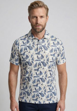 Homme avec une barbe claire portant un polo blanc à manches courtes avec un motif géométrique bleu et beige, debout devant un fond clair uni.