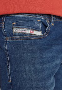 Jeans de denim de color azul profundo, con costuras naranjas, una etiqueta en el bolsillo trasero que dice "Diesel Industry" y un diseño clásico.