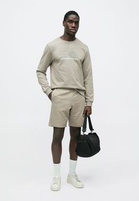 Bézs pulóver hímzett logóval, hozzáillő rövidnadrág, fehér zokni és bézs sportcipő; fekete táskát visel. Egyszerű, laza öltözet.