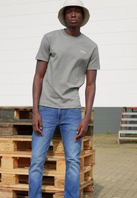 Camiseta de algodón gris con mangas cortas, con un pequeño logo blanco en el pecho, combinada con vaqueros de mezclilla azules. Palets de madera en el fondo.