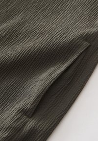 Tissu vert foncé avec une surface texturée et froissée et une couture visible le long du bord sur un fond blanc.