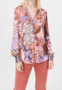 Femme portant un chemisier à manches longues à motifs floraux rose, violet et marron, associé à un pantalon en velours côtelé rose corail.
