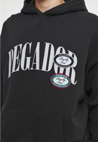 Sudadera negra con gran texto blanco "PEGADOR" y dos parches ovalados que dicen "Pegador Royalty Club Cold Hearted" en acentos en negro y verde azulado.