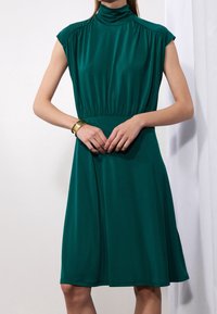 Kvinna iförd en ärmlös smaragdgrön knälång klänning med hög hals, händerna knäppta vid midjan och ett guldarmband på vänster handled.