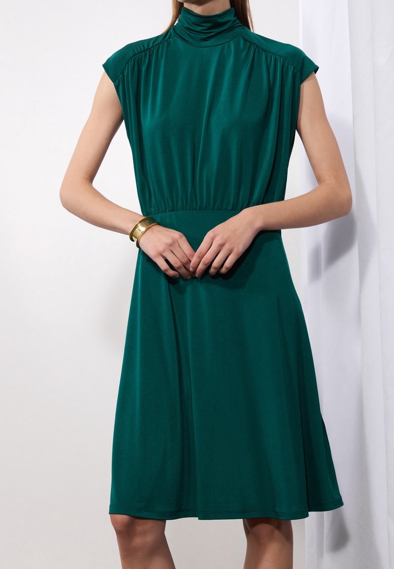 Kvinna iförd en ärmlös smaragdgrön knälång klänning med hög hals, händerna knäppta vid midjan och ett guldarmband på vänster handled.