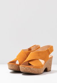 Sandali wedge gialli in suede con punta aperta, design incrociato, tacco in sughero e cinturino posteriore regolabile.
