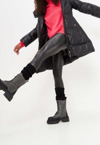 Grijze enkelboots met zwarte accenten, vetersluiting aan de voorkant en stevige rubberen zool. Model draagt zwarte leggings en een lange zwarte jas.