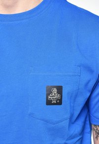 T-shirt blu realizzato in cotone con maniche corte, dotato di una tasca sul petto e un'etichetta nera rettangolare con un grafico di un cane, contrassegnato con la scritta "RefrigiWear".