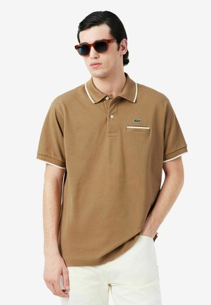 Man draagt een bruin poloshirt met korte mouwen en witte bies en zonnebril, staat met één hand in zijn zak tegen een effen achtergrond.