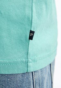 Tričko s krátkým rukávem v jemné modrozelené barvě, vyrobené z bavlny, s malou černou etiketou s logem na lemu a texturovaným žebrovaným vzorem.