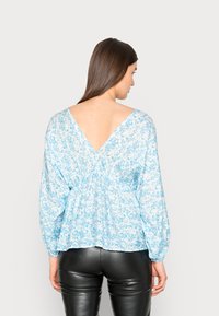 Blusa floral azul clara com decote em V nas costas, cintura elástica e mangas longas. O material é leve e fluido.