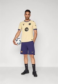 Kuldne jalgpallimängija lühikeste varrukatega särk tumesiniste detailide ja logodega; kombineeritud tumesiniste lühikeste pükste, mustade sokkide ja käes oleva jalgpalliga.