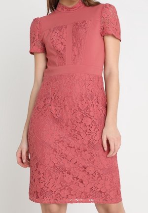 Mujer con un vestido de encaje color rosa polvo hasta la rodilla, con mangas cortas abullonadas, cuello alto y patrón floral en la falda y el corpiño.