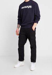 Sudadera azul marino con logotipo de marca en texto blanco, combinada con pantalones cargo negros con múltiples bolsillos y puños enrollados, usados con zapatillas blancas.