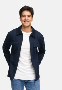 Marineblå lynlåsjakke med krave og to brystlommer. Bæret over en hvid skjorte, sammen med blå denimjeans. Glat tekstur.