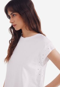 Hvid bomulds t-shirt med korte ærmer, prydet med metalliske nitter nær skuldrene. Glat tekstur og afslappet pasform.