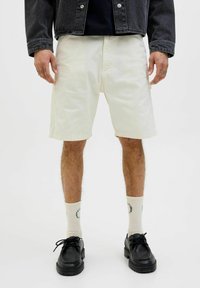 Shorts blancs décontractés en tissu léger, arrivant aux genoux, avec des poches latérales. Associés à des chaussures noires et des chaussettes crème avec un texte imprimé.